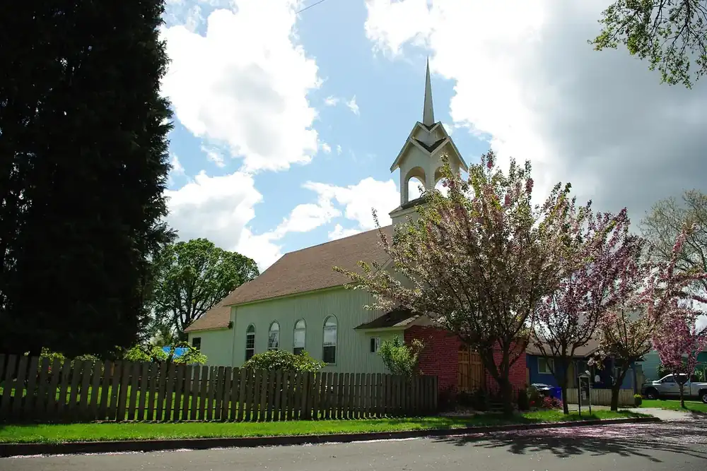 White church with a tall steeple behind a wooden fence and flowering trees in Cornelius