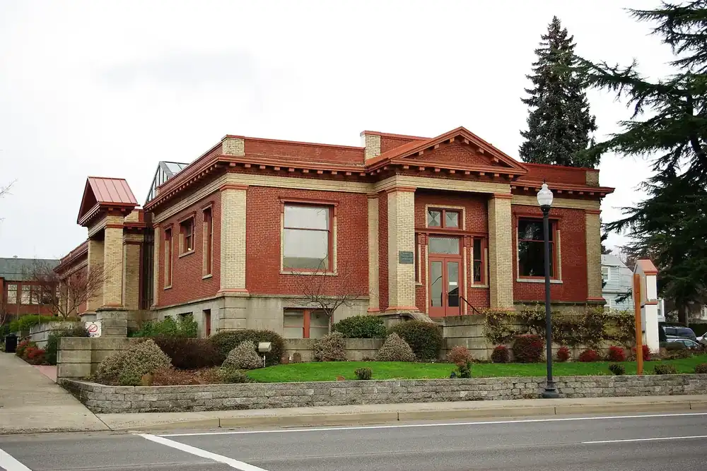 Red-brick Carnegie library with tan columns and landscaped beds along a street in Newberg, Oregon